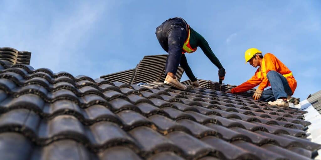 Two workers in safety gear installing tiles on a steep residential roof.