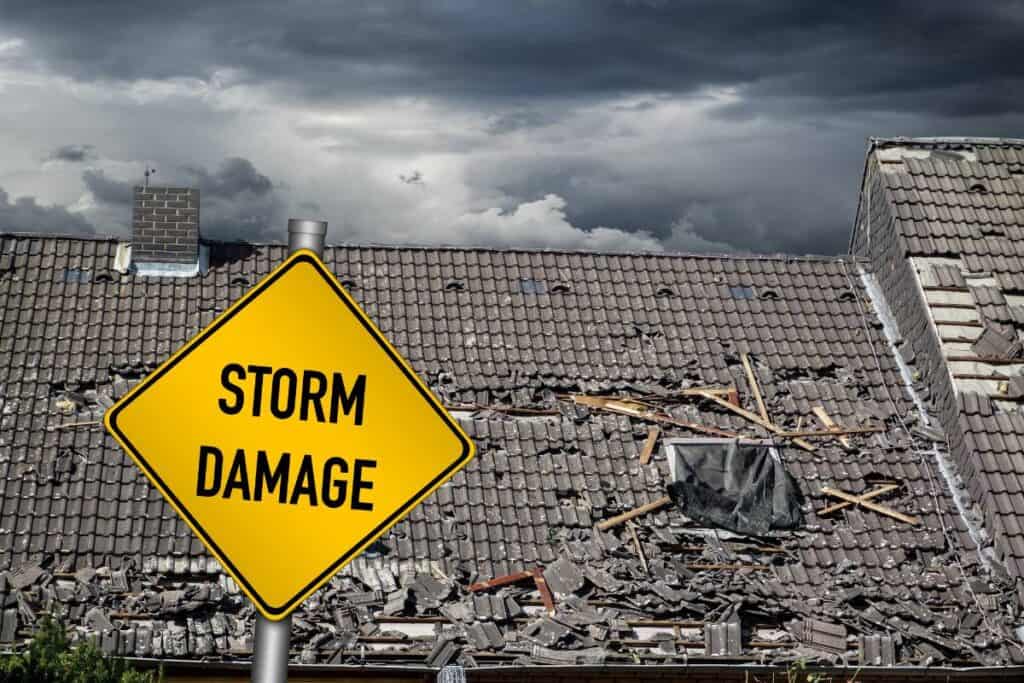 A "STORM DAMAGE" road sign in front of a severely damaged home in need of roof repair.